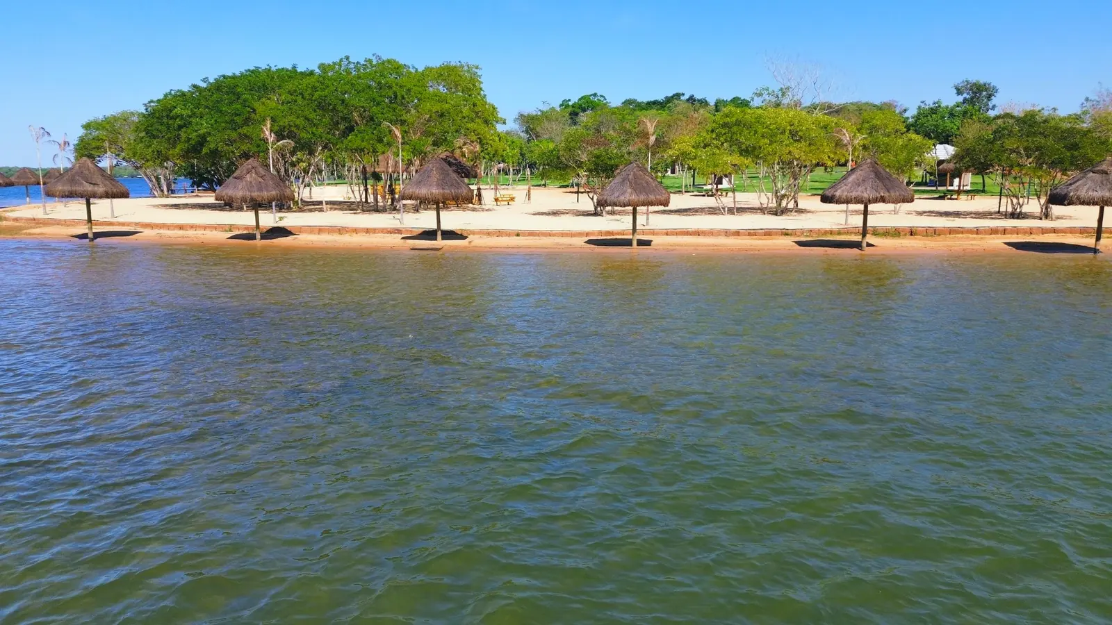 Orla do Quinta do Lago vista da água, com quiosques de palha na areia e árvores atrás